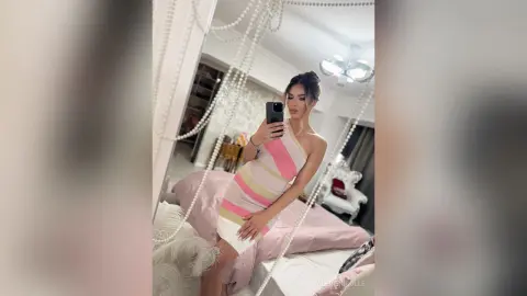 Young woman in a pink striped bodycon dress taking a mirror selfie in a bright, modern bedroom.