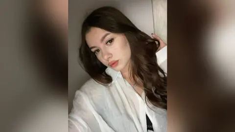 Young woman with long wavy brown hair wearing a white linen shirt in a soft, natural indoor setting.