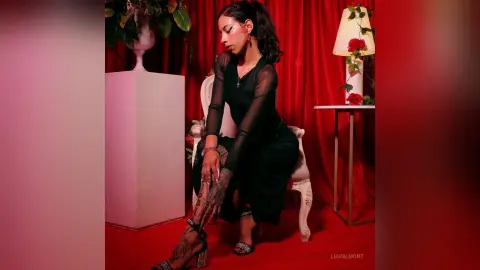 LiaValmont sits in a white chair wearing a sheer black long-sleeve top and black pants, posing against a red curtain background.