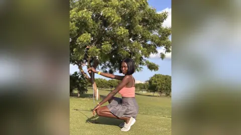 Young woman in a pink tank top and grey skirt playing golf on a sunny green field.