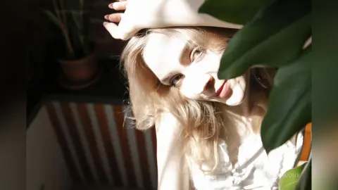 Blonde woman in a white summer dress relaxing in sunlit indoor greenery with a peaceful mood.