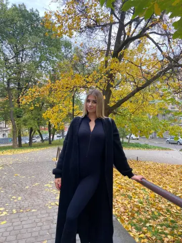 Blonde woman in a black jumpsuit and long cardigan posing in a park with autumn leaves.