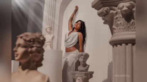 Elegant woman in a white flowing dress posing amidst classical marble statues in a dreamy, ethereal setting.