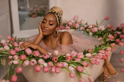 Glamorous Black woman in a pink tulle gown relaxing in a bathtub filled with pink tulips for a dreamy photoshoot.