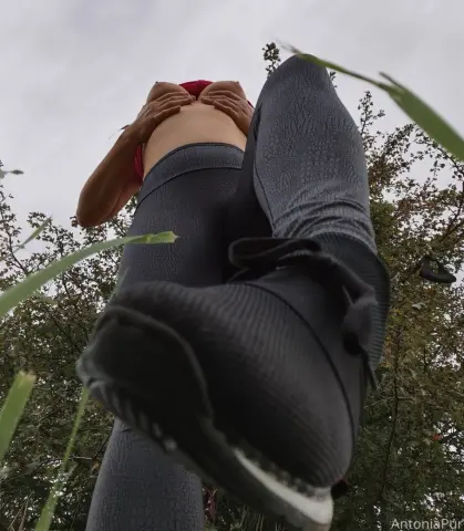 AntoniaPur28 poses outdoors in black leggings and a red top, using both hands to touch and lift her pussy while looking down at the camera.