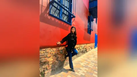 Young woman in a black jacket and jeans leaning against a vibrant red wall in a sunny urban setting.