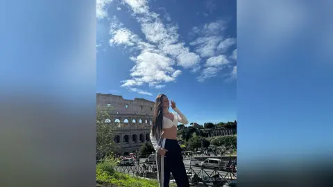 Young woman in white crop top and black leggings posing against a blue sky near the Roman Colosseum.