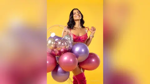 Smiling woman in a pink corset holding colorful balloons against a bright yellow background.