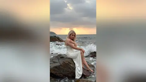 Blonde woman in a white maxi dress sitting on rocks by the ocean during a moody sunset.