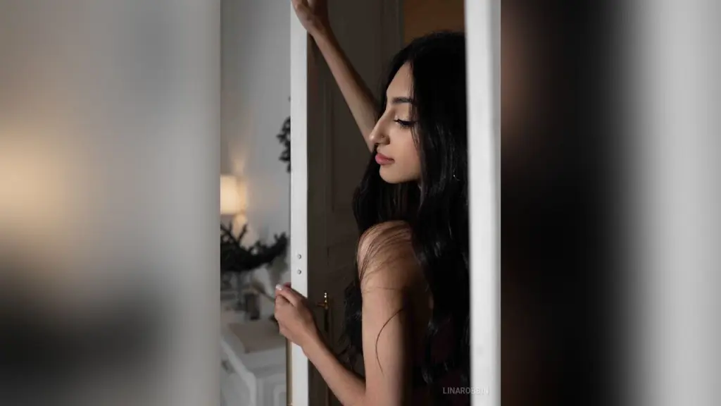 LinaRobbin — Elegant woman in a black dress standing by a doorway in a soft, moody indoor setting.