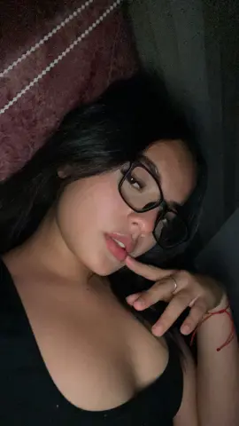 Katalinax_Rose wearing black glasses and a black top, posing in bed with her finger to her lips.