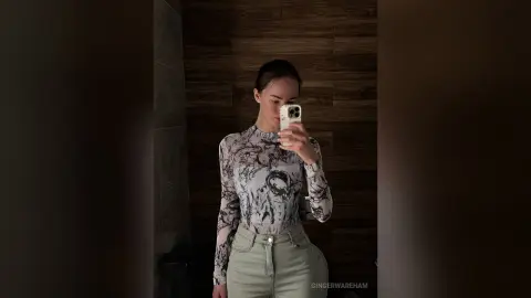 Young woman in a floral print turtleneck and light jeans taking a moody mirror selfie in a dark room.