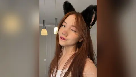 Smiling young woman with long brown hair wearing black cat ears in a cozy indoor setting.