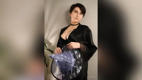Young woman in a black satin robe and choker holding a sparkly blue witch hat in a moody indoor setting.