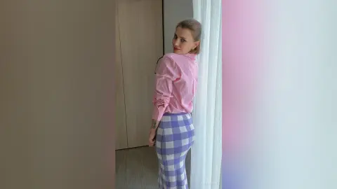 Blonde woman in pink blouse and purple checkered skirt posing by a window in a bright room.