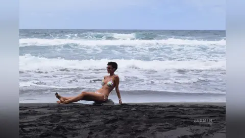 Smiling woman in a floral bikini relaxing on a dark volcanic sand beach by the ocean waves.