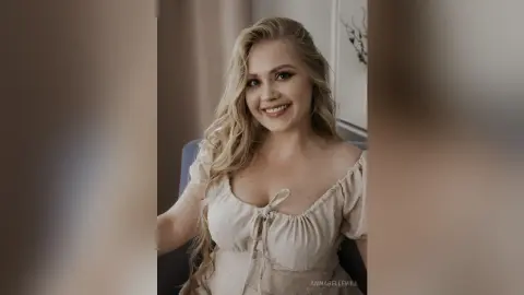 Smiling blonde woman in a white puff-sleeve summer dress sitting in a bright, cozy indoor setting.