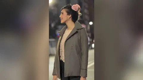 HaleyLyon smiles in profile wearing a grey coat and a light lace top with a pink flower in her hair while standing outside at night.