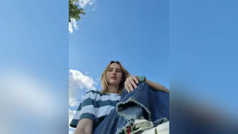 LisetteMaupredi poses outdoors against a blue sky, wearing a striped t-shirt and holding up a pair of denim jeans.