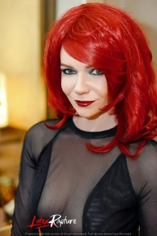 LatexRapture has bright red hair and wears a black sheer mesh top while looking at the camera.