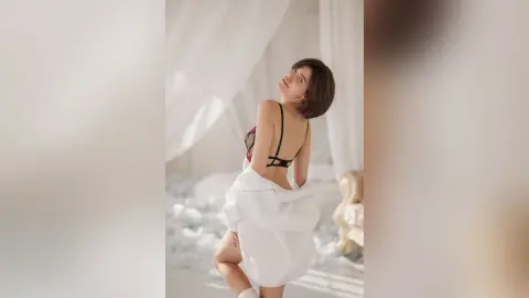 Elegant woman in black lingerie and white robe posing in a bright, airy minimalist bedroom.
