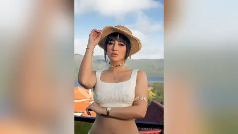 Stylish woman in a white ribbed crop top and straw hat posing outdoors against a scenic summer landscape.