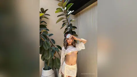Smiling woman in a white lace boho outfit posing next to indoor plants in a bright, airy room.