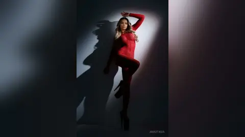 Anastasia poses in a red long-sleeve bodysuit with her arm raised against a dark background.