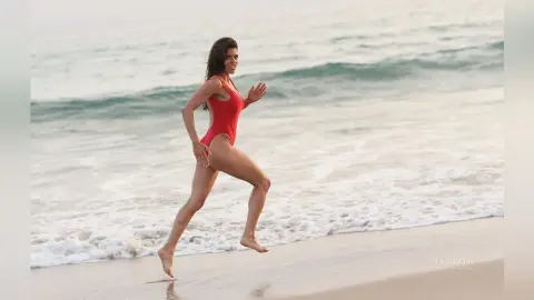 Fit woman in a red one-piece swimsuit running joyfully along a sunny beach with ocean waves.