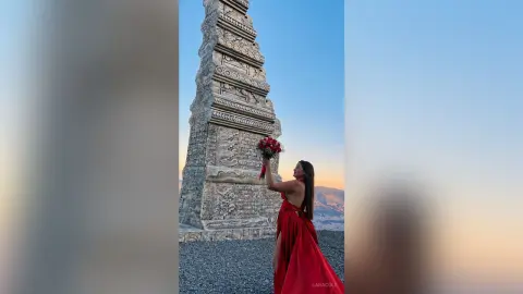 LaraCole stands outdoors in a long red dress, holding a bouquet of red flowers next to a tall, carved stone monument under a clear blue sky.