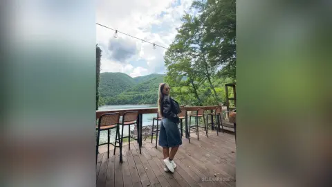 SelenaRusse stands on a wooden deck overlooking a lake and green mountains, wearing a black leather jacket and a denim skirt.