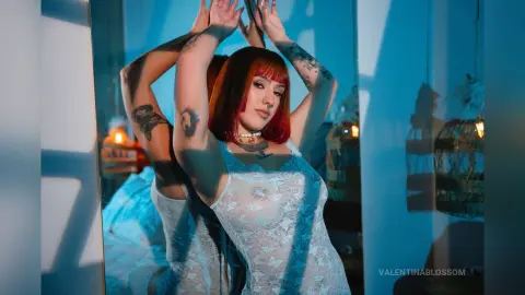 Tattooed woman in white lace lingerie posing in moody blue sunlight by a mirror.