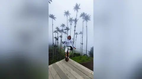 AmelieLautrec stands on a wooden walkway surrounded by tall palm trees in a foggy outdoor setting, wearing a white long-sleeve top and white shorts.