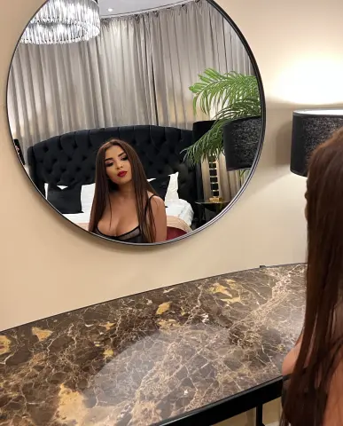 Glamorous woman in a black top reflected in a round mirror within a luxury modern bedroom setting.