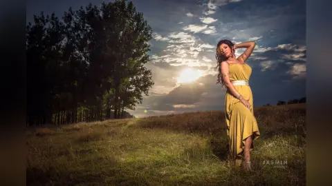 MissyJolie poses in a long yellow dress against a bright, cloudy sky in an open grassy field.