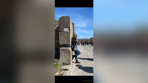 Woman in black turtleneck and jeans standing by ancient stone ruins under a bright blue sky.