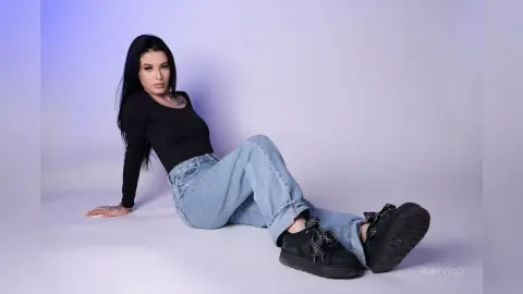 Young woman in black long-sleeve shirt and light blue jeans sitting on a white studio background.