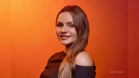 MilaVogue smiles while looking at the camera, wearing a black off-the-shoulder top against a bright orange background.