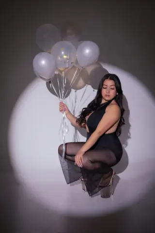 Elegant woman in a black bodysuit and sheer skirt holding white balloons in a moody studio setting.