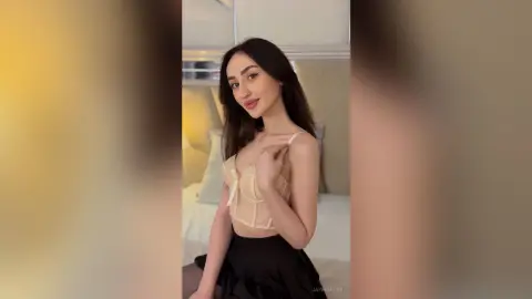 Young woman in a beige corset top and black skirt posing in a bright, cozy bedroom setting.