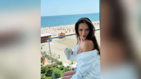 Beautiful woman in a white robe sipping coffee on a sunny balcony overlooking a tropical beach.