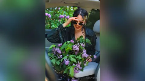 Smiling woman in black leather jacket holding a large bouquet of purple lilacs in a car seat.