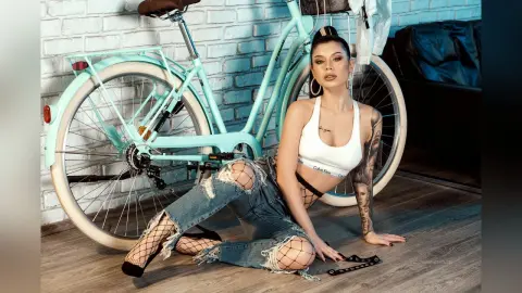 Stylish woman in white tank top and ripped jeans sitting by a vintage light blue bicycle in a bright room.