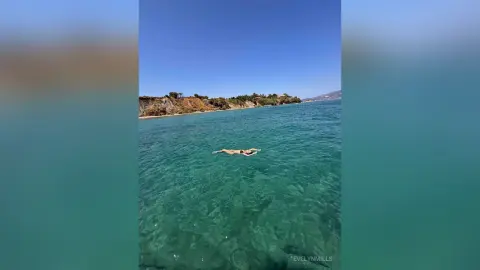 EvelynMills floats naked in clear turquoise ocean water near a rocky coastline under a blue sky.