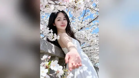 Smiling young woman in a white summer dress posing under blooming cherry blossom trees in bright daylight.
