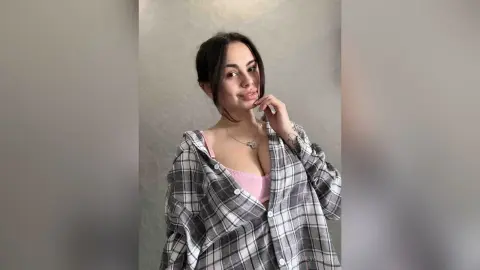 Smiling young woman in a pink camisole and plaid shirt posing against a neutral background.