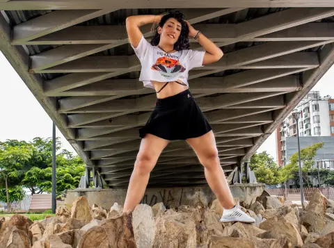 Young woman in white graphic t-shirt and black mini skirt posing confidently under a steel bridge structure.