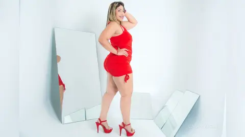 Curvy woman in a red mini dress and high heels posing confidently in a bright white studio with mirrors.