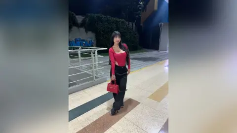 Stylish woman in a red long-sleeve top and black trousers holding a red handbag on a city sidewalk at night.