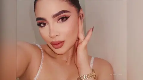 Woman with glamorous winged eyeliner and nude makeup wearing a white lace camisole in a soft indoor setting.